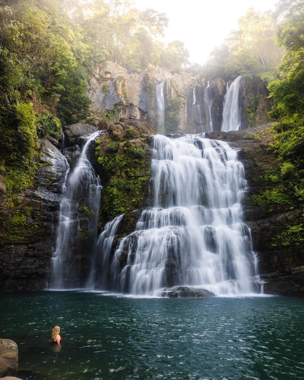 Mujer en un traje de baño rojo disfrutando de una cascada rodeada de vegetación exuberante y un lago de agua cristalina. La imagen captura la belleza natural de un entorno tropical con múltiples caídas de agua.