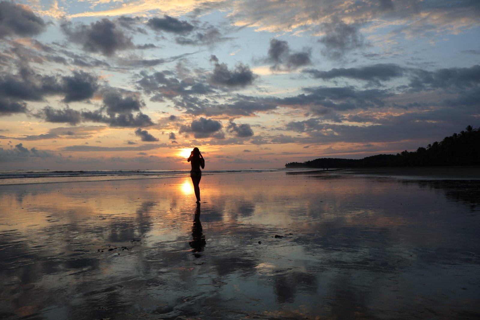 Silhouette of a person standing on a reflective beach at sunset, with colorful clouds and gentle waves in the background.