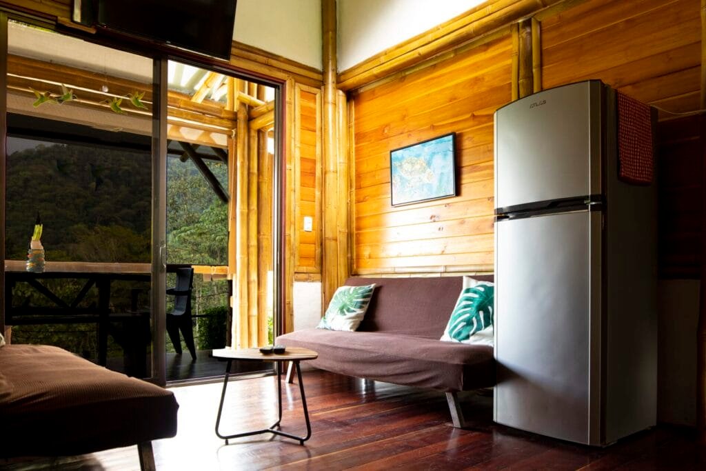 Interior de una cabaña moderna con paredes de madera, un sofá cómodo, una mesa pequeña y una nevera. La luz natural entra a través de grandes ventanales, creando un ambiente acogedor y relajante.