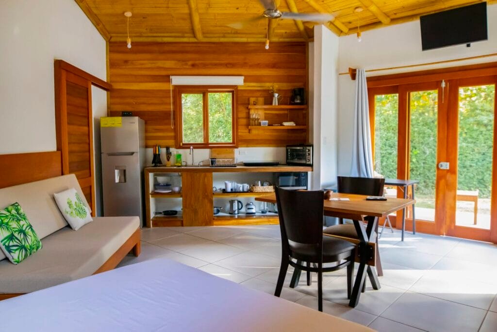 Interior de una cabaña moderna con cocina equipada, sofá cómodo y mesa de comedor. Ventanas grandes que permiten la entrada de luz natural y acceso a un jardín. Decoración con elementos naturales y plantas. Ideal para vacaciones relajantes.
