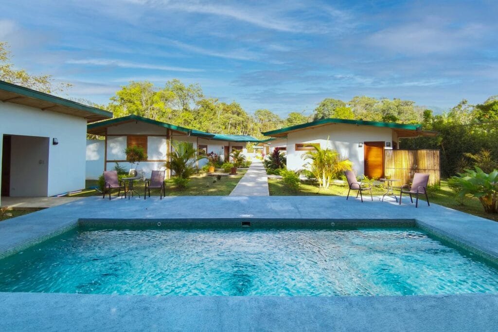Vista de un complejo turístico con cabañas rodeadas de jardines verdes y una piscina en primer plano, bajo un cielo azul despejado. Ideal para vacaciones relajantes.