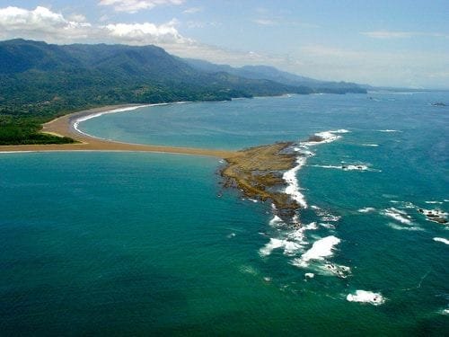Aerial view of a stunning coastal landscape featuring a sandy beach, lush mountains in the background, and waves crashing against rocky outcrops. Ideal for showcasing natural beauty and outdoor adventure destinations.