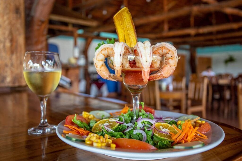 Delicious shrimp cocktail served in a glass, garnished with a fried plantain, alongside a colorful fresh salad featuring lettuce, tomatoes, carrots, cucumbers, and corn, with a glass of white wine in a rustic restaurant setting.