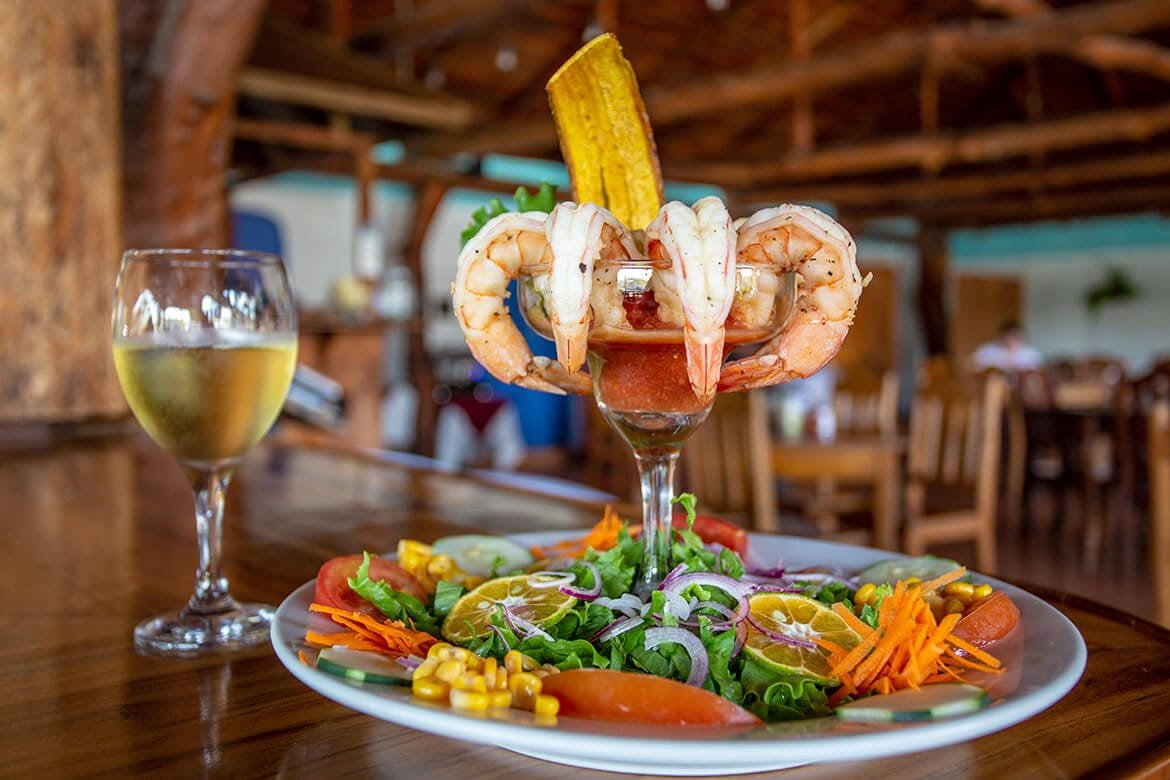 Delicious shrimp cocktail served in a glass, garnished with a fried plantain, alongside a colorful fresh salad featuring lettuce, tomatoes, carrots, cucumbers, and corn, with a glass of white wine in a rustic restaurant setting.