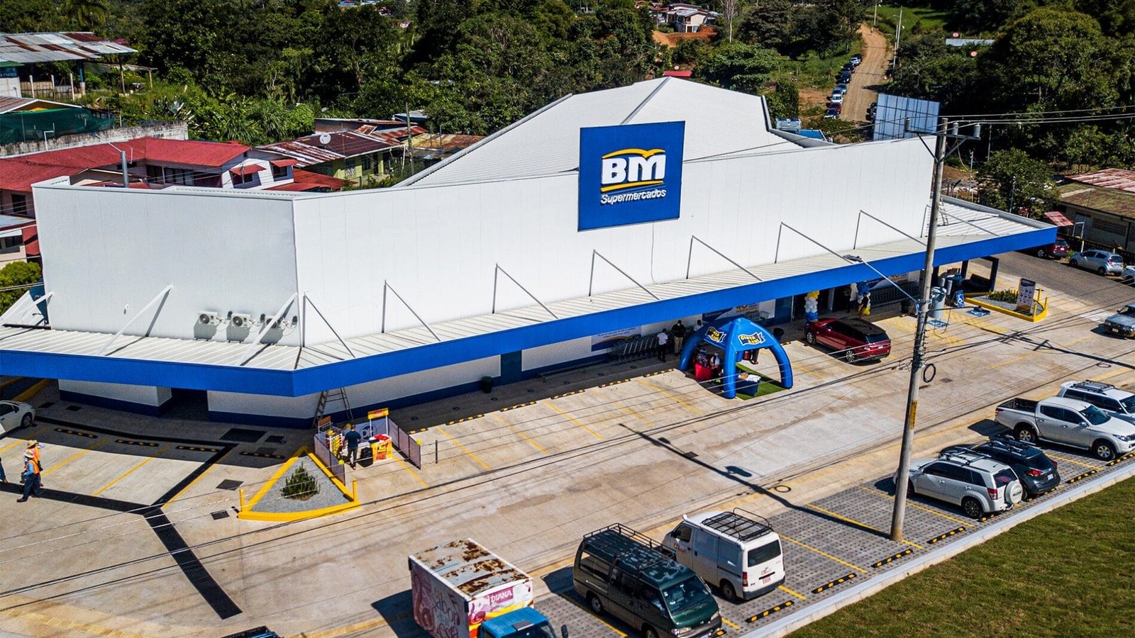 Aerial view of BM Supermercados, showcasing the modern storefront, parking lot filled with vehicles, and surrounding greenery, highlighting its location in a residential area.