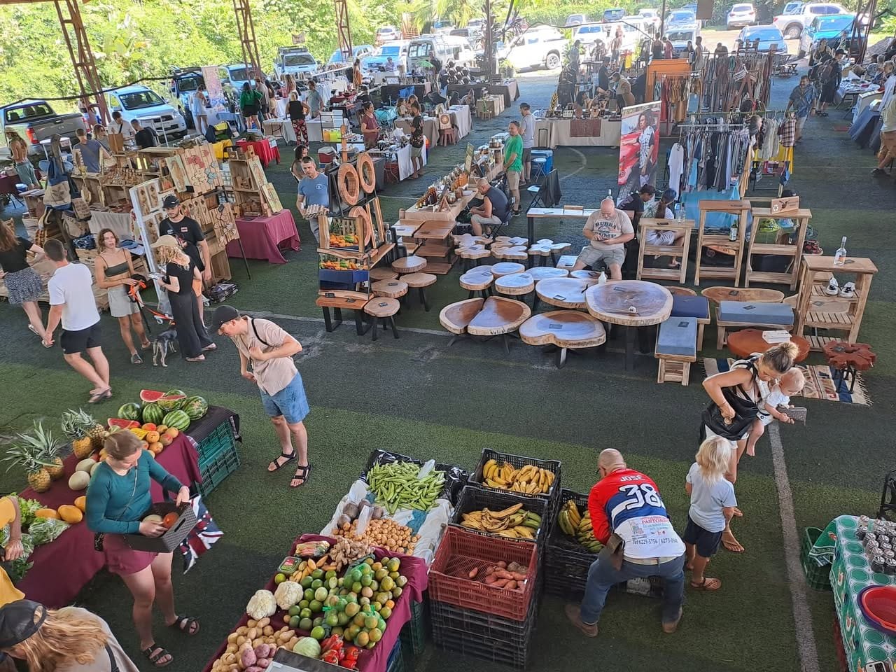 Mercado local en un espacio cubierto, con vendedores ofreciendo frutas frescas, vegetales y productos artesanales. Visitantes interactuando y comprando, rodeados de mesas de madera y una variedad de productos. Ambiente animado y colorido.