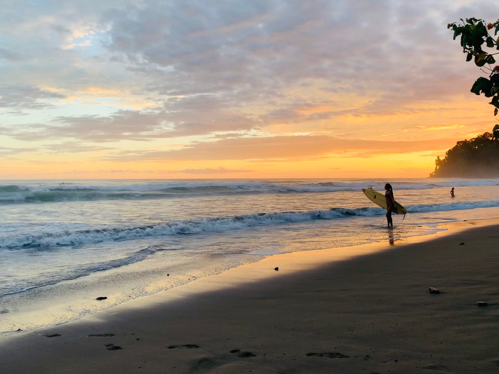 Surfer walking along a sandy beach at sunset, with gentle waves lapping at the shore and a colorful sky filled with clouds.