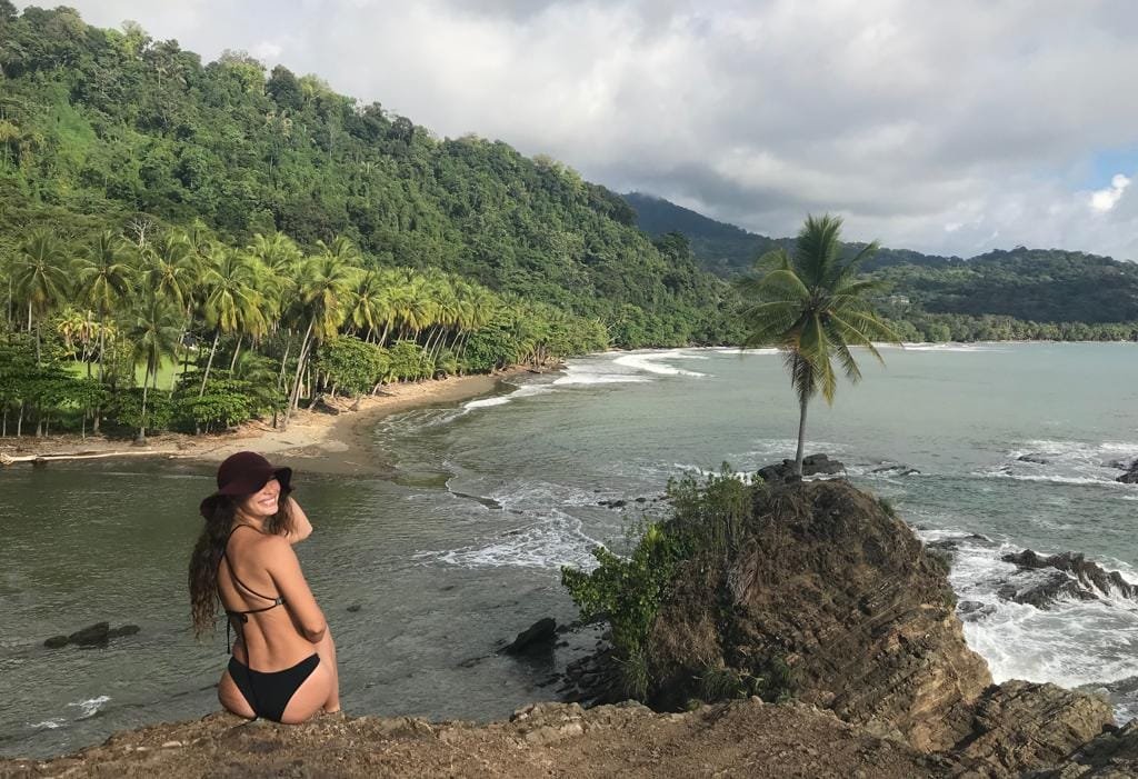 Mujer sonriente con un sombrero en la playa, rodeada de palmeras y montañas verdes, disfrutando de la vista al mar en un paisaje tropical.