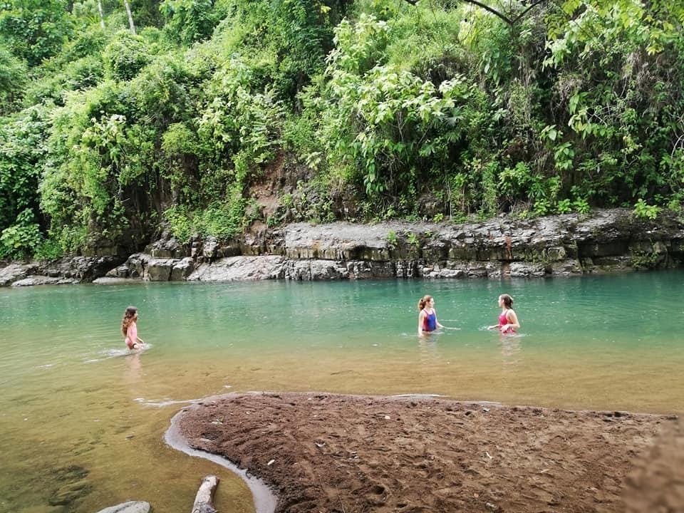 Three children playing in a natural swimming hole surrounded by lush greenery and rocky terrain, enjoying a sunny day outdoors.