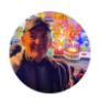Hombre sonriendo en un entorno festivo iluminado, con coloridas decoraciones de fondo.
