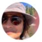 Retrato de una persona con gafas de sol y sombrero en un entorno natural, con árboles y un lago de fondo.