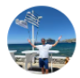 Hombre sonriente de pie junto al mar, con brazos abiertos y fondo de paisaje costero en un día soleado.